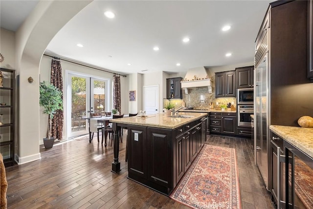 kitchen with light stone counters, dark wood-style floors, premium range hood, arched walkways, and decorative backsplash