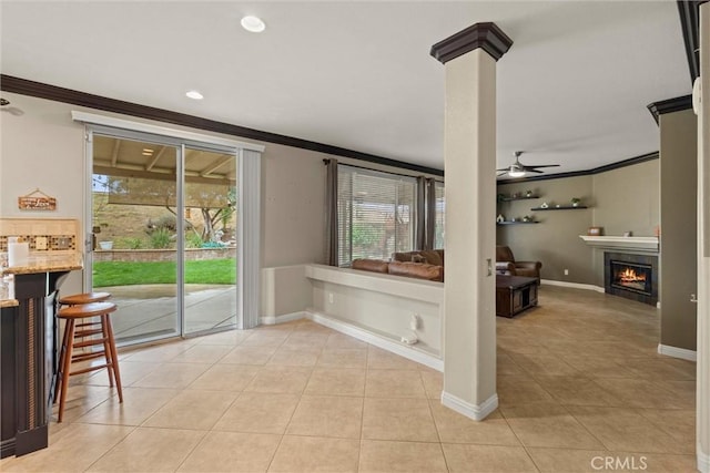 interior space with a warm lit fireplace, tile patterned flooring, a ceiling fan, baseboards, and crown molding