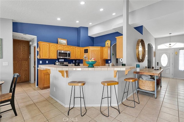 kitchen with a breakfast bar, tile countertops, light tile patterned floors, appliances with stainless steel finishes, and a peninsula