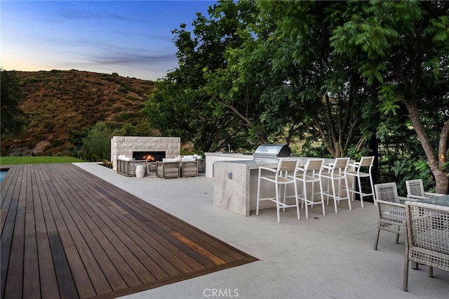deck at dusk featuring exterior kitchen, an outdoor stone fireplace, a patio area, and grilling area