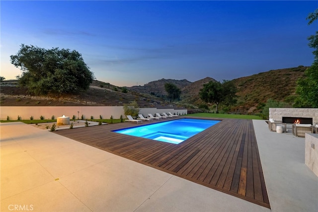 pool at dusk with a patio, an outdoor stone fireplace, a mountain view, and a fenced in pool