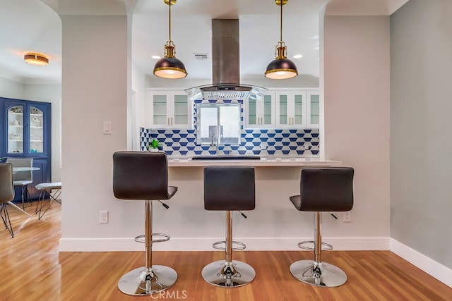 kitchen featuring light wood finished floors, decorative backsplash, glass insert cabinets, a breakfast bar area, and island exhaust hood