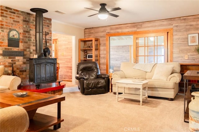 carpeted living room featuring ceiling fan, wooden walls, visible vents, a wood stove, and crown molding