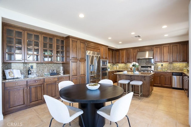 kitchen featuring a center island, tasteful backsplash, glass insert cabinets, built in appliances, and under cabinet range hood