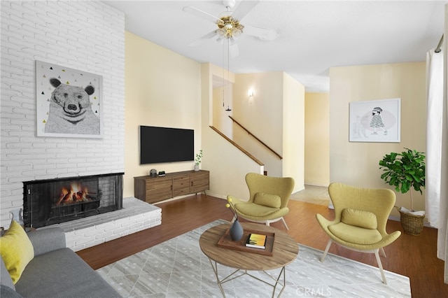 living area with ceiling fan, wood finished floors, baseboards, stairs, and a brick fireplace