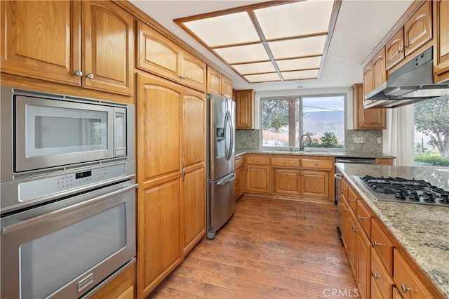 kitchen featuring under cabinet range hood, wood finished floors, a sink, appliances with stainless steel finishes, and light stone countertops