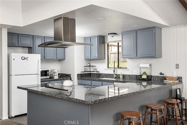 kitchen featuring island range hood, dark stone counters, stainless steel microwave, freestanding refrigerator, and a peninsula