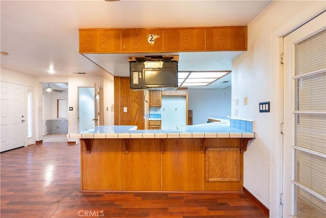 kitchen with a peninsula, dark wood-style floors, tile counters, and brown cabinetry