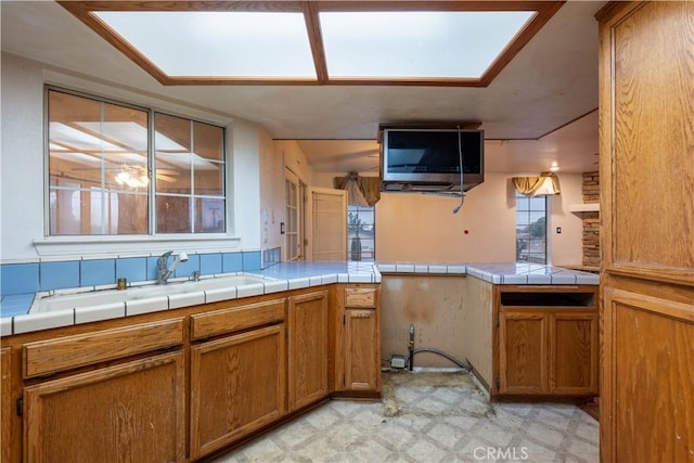 kitchen with tile countertops, brown cabinets, a peninsula, light floors, and a sink