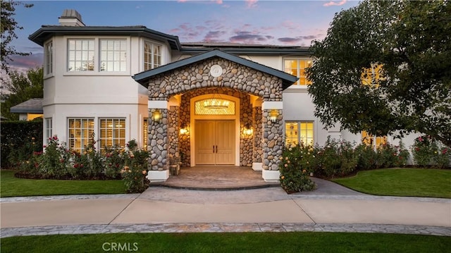 exterior entry at dusk featuring a yard, a chimney, and stucco siding