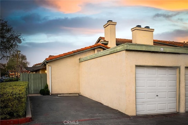 property exterior at dusk featuring a garage, a tile roof, fence, and stucco siding