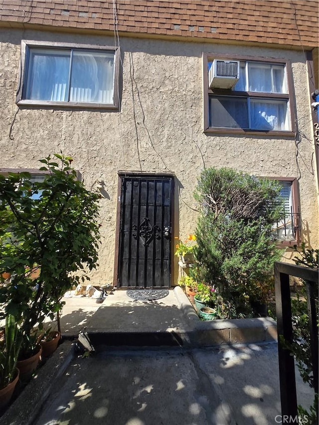 view of exterior entry with a shingled roof, an AC wall unit, and stucco siding