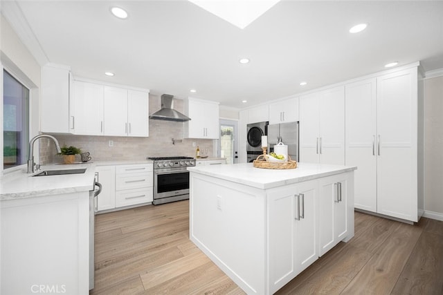 kitchen featuring stainless steel appliances, stacked washer and dryer, a sink, light wood-type flooring, and wall chimney exhaust hood