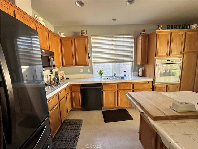 kitchen with tile counters, recessed lighting, brown cabinetry, black appliances, and a sink