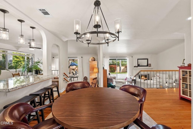 dining room with arched walkways, a warm lit fireplace, ceiling fan with notable chandelier, wood finished floors, and visible vents