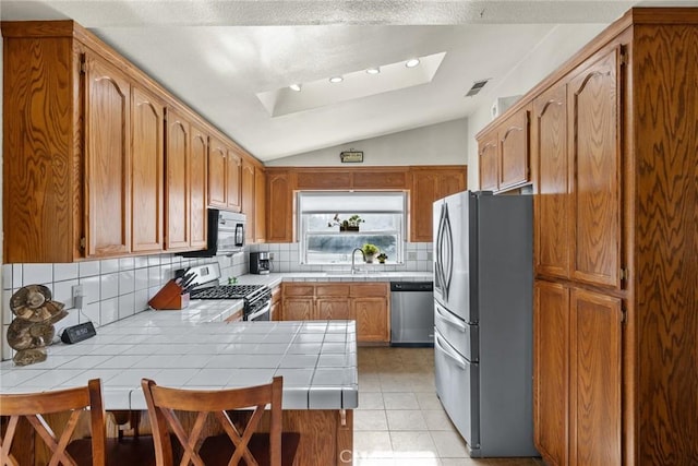 kitchen with tile countertops, vaulted ceiling with skylight, a peninsula, a sink, and appliances with stainless steel finishes