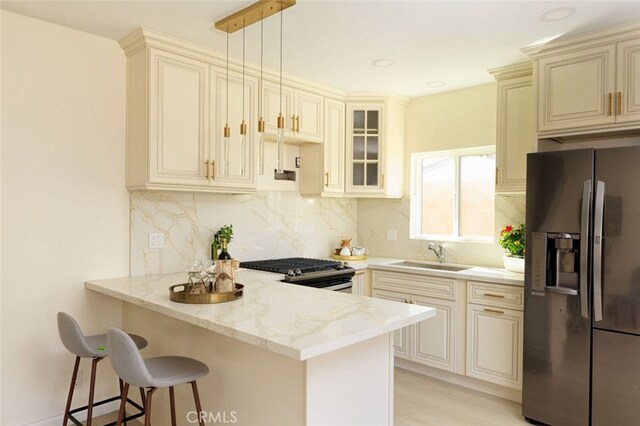 kitchen with light stone countertops, a peninsula, a sink, cream cabinetry, and appliances with stainless steel finishes