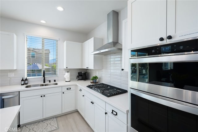 kitchen featuring stainless steel appliances, a sink, white cabinetry, light countertops, and wall chimney range hood