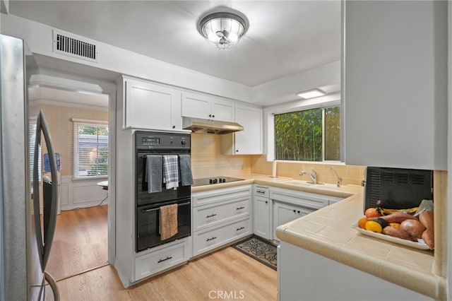 kitchen with light wood finished floors, visible vents, tile counters, under cabinet range hood, and black appliances