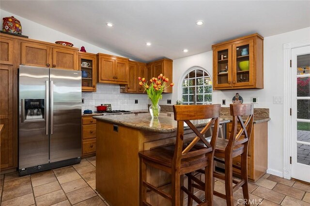 kitchen featuring tasteful backsplash, dark stone counters, brown cabinetry, stainless steel fridge with ice dispenser, and vaulted ceiling