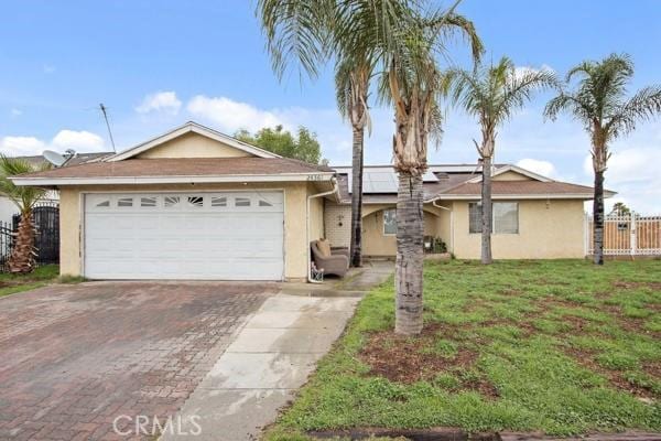 ranch-style home featuring stucco siding, decorative driveway, roof mounted solar panels, fence, and a front yard