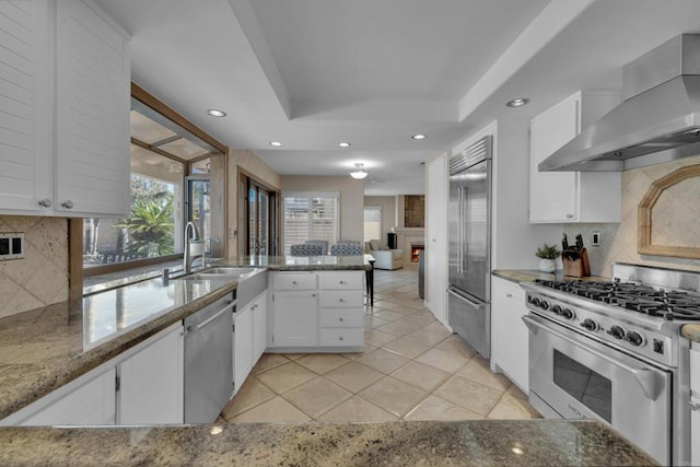 kitchen with a large fireplace, a raised ceiling, stainless steel appliances, wall chimney range hood, and a sink