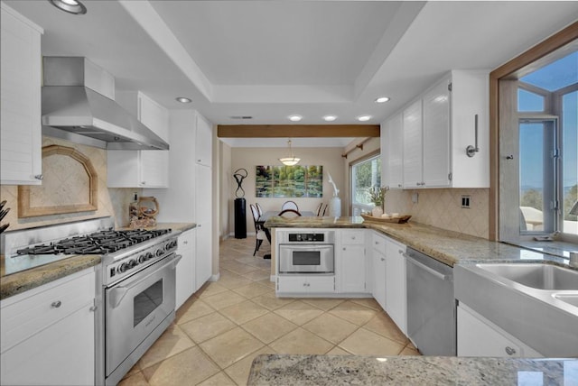 kitchen with white cabinets, appliances with stainless steel finishes, a peninsula, a tray ceiling, and wall chimney range hood