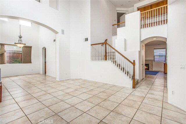 tiled entrance foyer featuring a warm lit fireplace, visible vents, arched walkways, a towering ceiling, and stairway