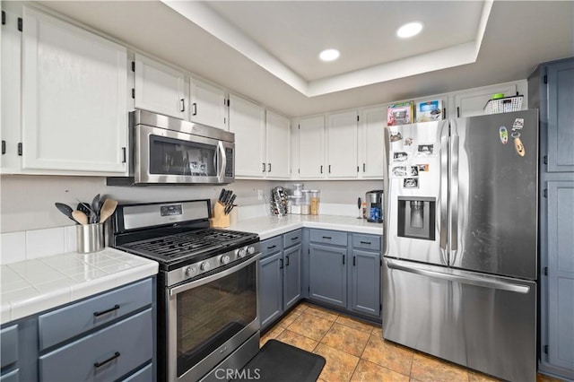 kitchen with tile counters, appliances with stainless steel finishes, white cabinets, and a tray ceiling