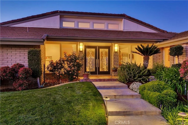 entrance to property featuring french doors, a tiled roof, covered porch, a yard, and brick siding