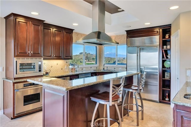 kitchen with light stone counters, island exhaust hood, a sink, built in appliances, and a kitchen breakfast bar