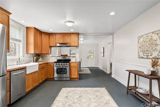 kitchen with brown cabinetry, appliances with stainless steel finishes, a sink, light countertops, and backsplash