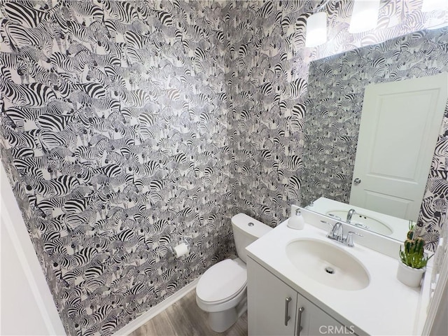 bathroom with wallpapered walls, wood finished floors, toilet, and vanity