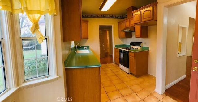 kitchen with baseboards, brown cabinetry, gas range, under cabinet range hood, and a sink