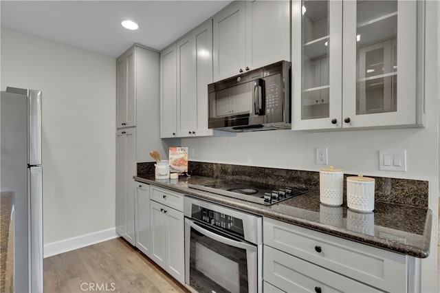 kitchen with stainless steel appliances, baseboards, light wood-style floors, dark stone counters, and glass insert cabinets