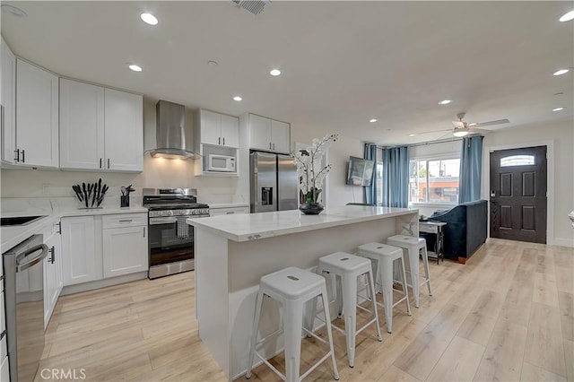 kitchen featuring light wood finished floors, a kitchen island, appliances with stainless steel finishes, a breakfast bar, and wall chimney range hood