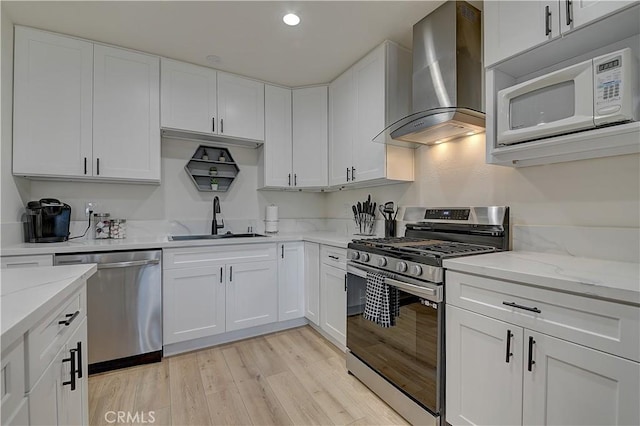 kitchen featuring appliances with stainless steel finishes, light wood-style floors, white cabinets, a sink, and wall chimney exhaust hood