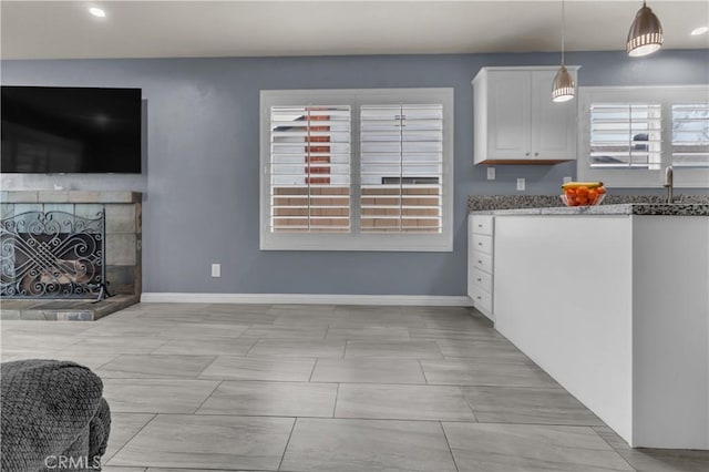 kitchen with a fireplace with raised hearth, baseboards, dark stone countertops, hanging light fixtures, and white cabinetry