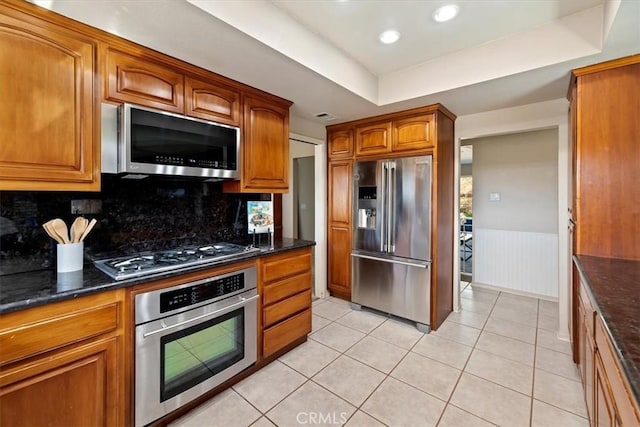 kitchen featuring light tile patterned floors, stainless steel appliances, wainscoting, brown cabinets, and a tray ceiling