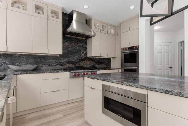 kitchen featuring light wood-type flooring, dark stone counters, appliances with stainless steel finishes, wall chimney exhaust hood, and decorative backsplash