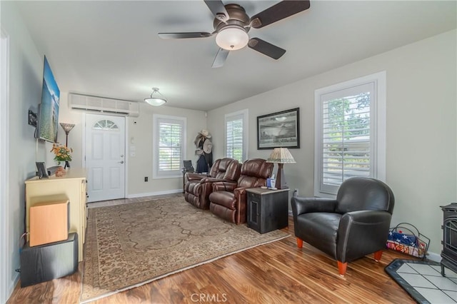 living room with a wall unit AC, a wood stove, ceiling fan, wood finished floors, and baseboards