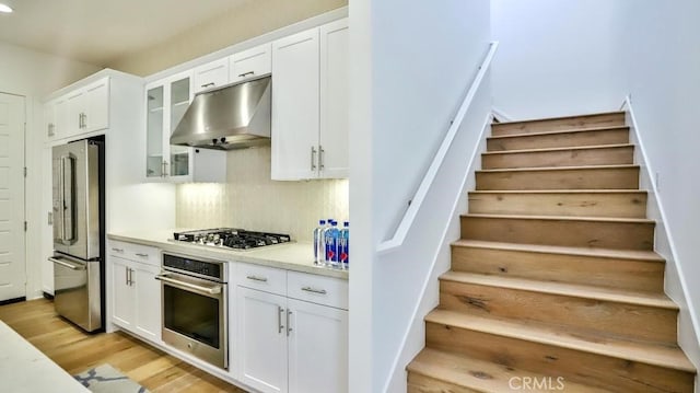 kitchen with stainless steel appliances, glass insert cabinets, white cabinetry, light wood-type flooring, and under cabinet range hood