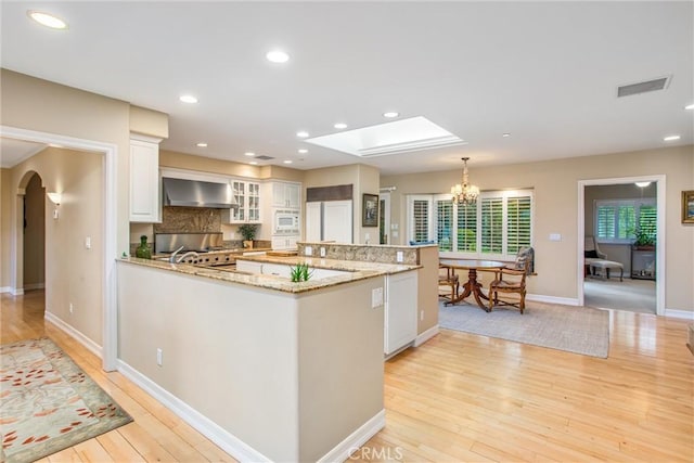 kitchen with arched walkways, white cabinets, light wood-style floors, white microwave, and wall chimney range hood