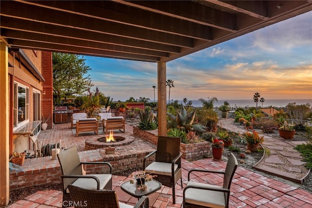 patio terrace at dusk with area for grilling, a fire pit, and fence