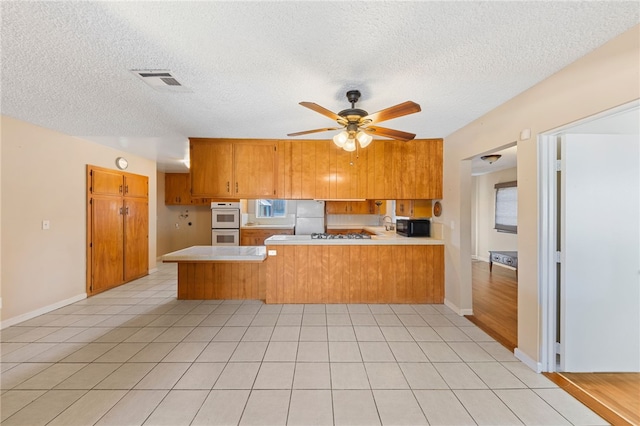 kitchen with a peninsula, white appliances, visible vents, light countertops, and brown cabinetry