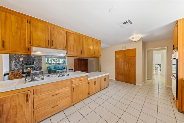 kitchen featuring light tile patterned floors, visible vents, a stone fireplace, under cabinet range hood, and white gas cooktop