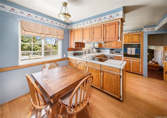 kitchen with brown cabinetry, tile countertops, a peninsula, light wood-type flooring, and black appliances