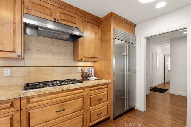 kitchen with under cabinet range hood, decorative backsplash, brown cabinets, appliances with stainless steel finishes, and wood finished floors