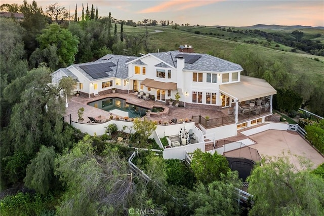 back of house at dusk with a fenced backyard, a chimney, an in ground hot tub, a patio area, and roof mounted solar panels