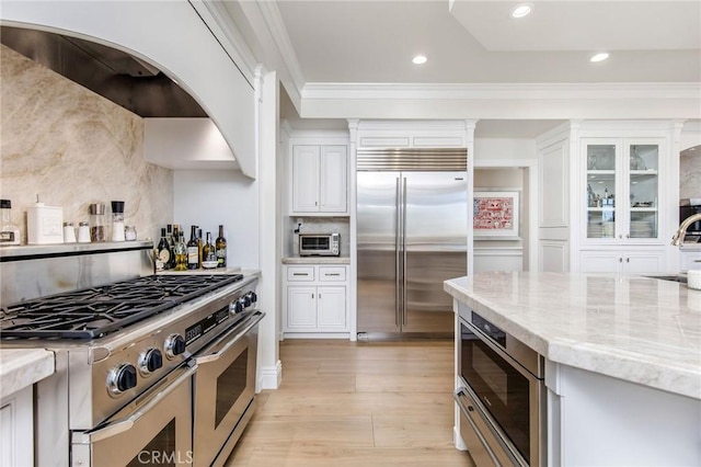 kitchen with built in appliances, light wood-style floors, white cabinets, backsplash, and light stone countertops